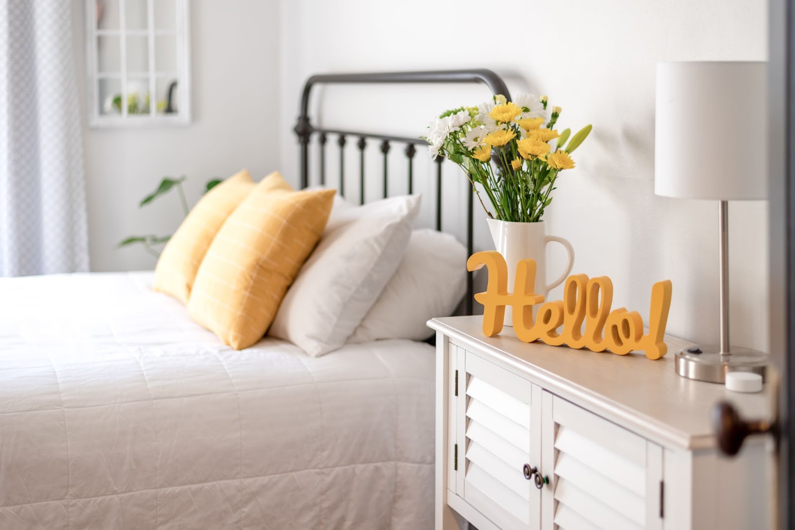 A cozy bedroom with a bed, yellow pillows, and a nightstand featuring flowers and a Hello sign.