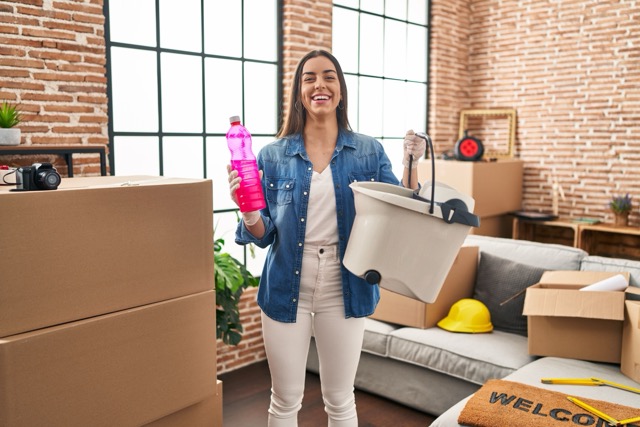 A smiling woman holding a pink cleaning bottle and a mop bucket while standing in a room filled with moving boxes.