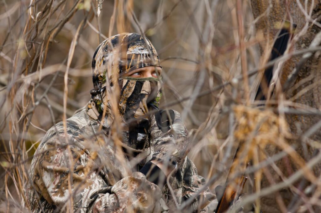 Hunter in camouflage hiding in bushes with a rifle.