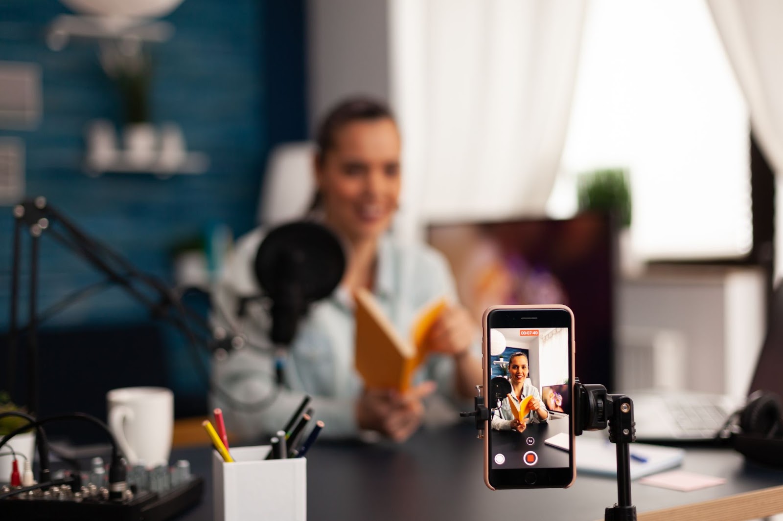 A person recording a video with a smartphone on a stand, holding a book, with a microphone and desk items in the background.