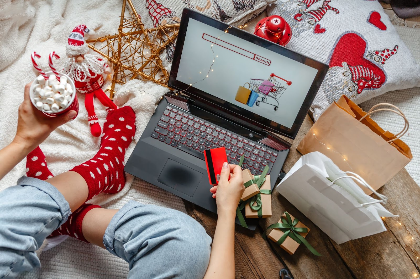 A person with red polka-dot socks holds a credit card and a cup of marshmallows while shopping online on a laptop surrounded by Christmas decorations and gift bags.