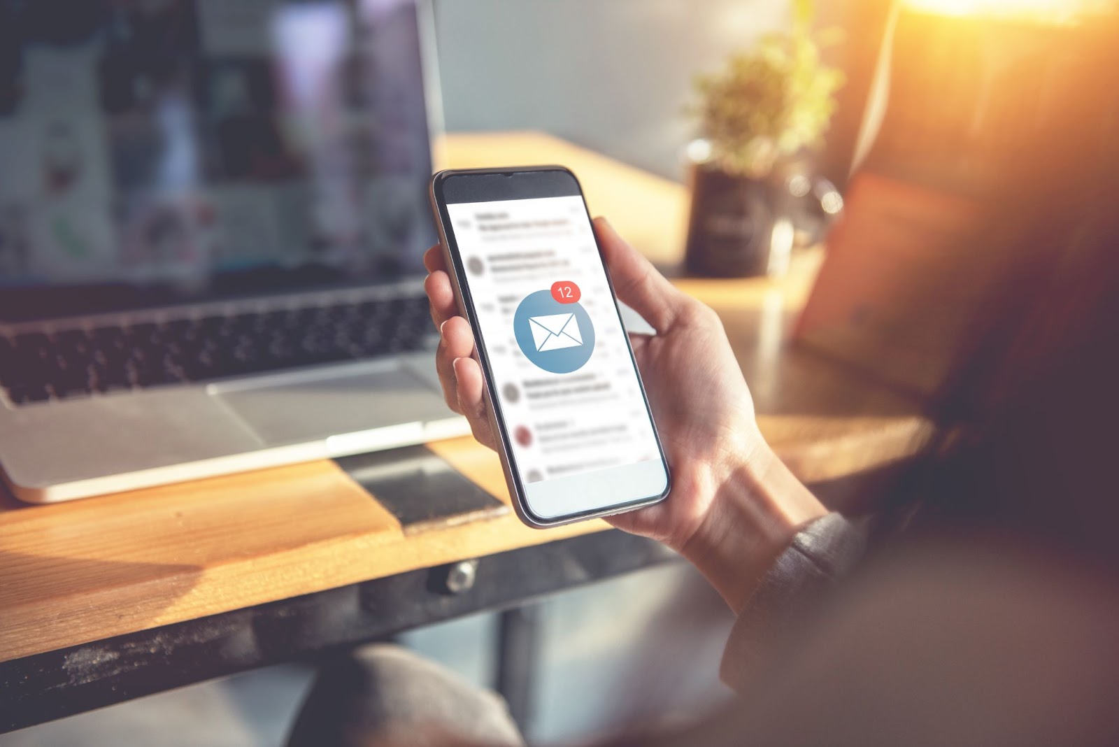 Person holding a smartphone with email notifications, working at a desk with a laptop.