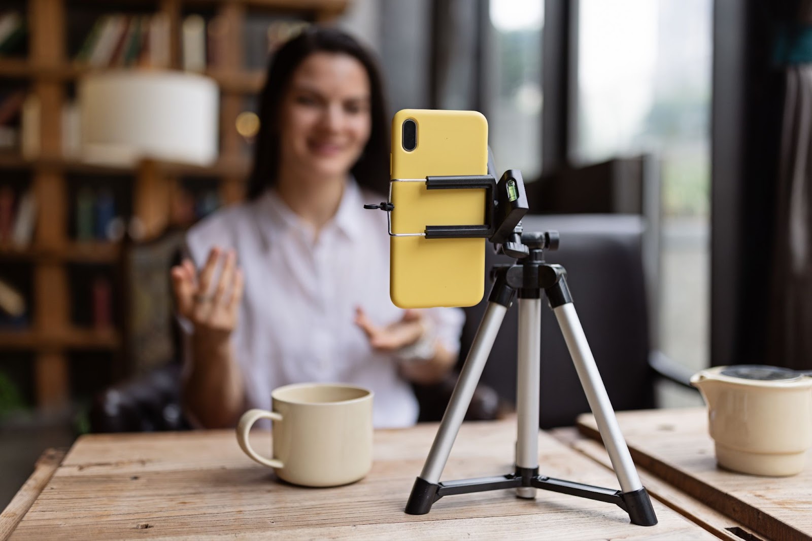 Woman recording video with phone on tripod in cozy setting with bookshelves and coffee mug.